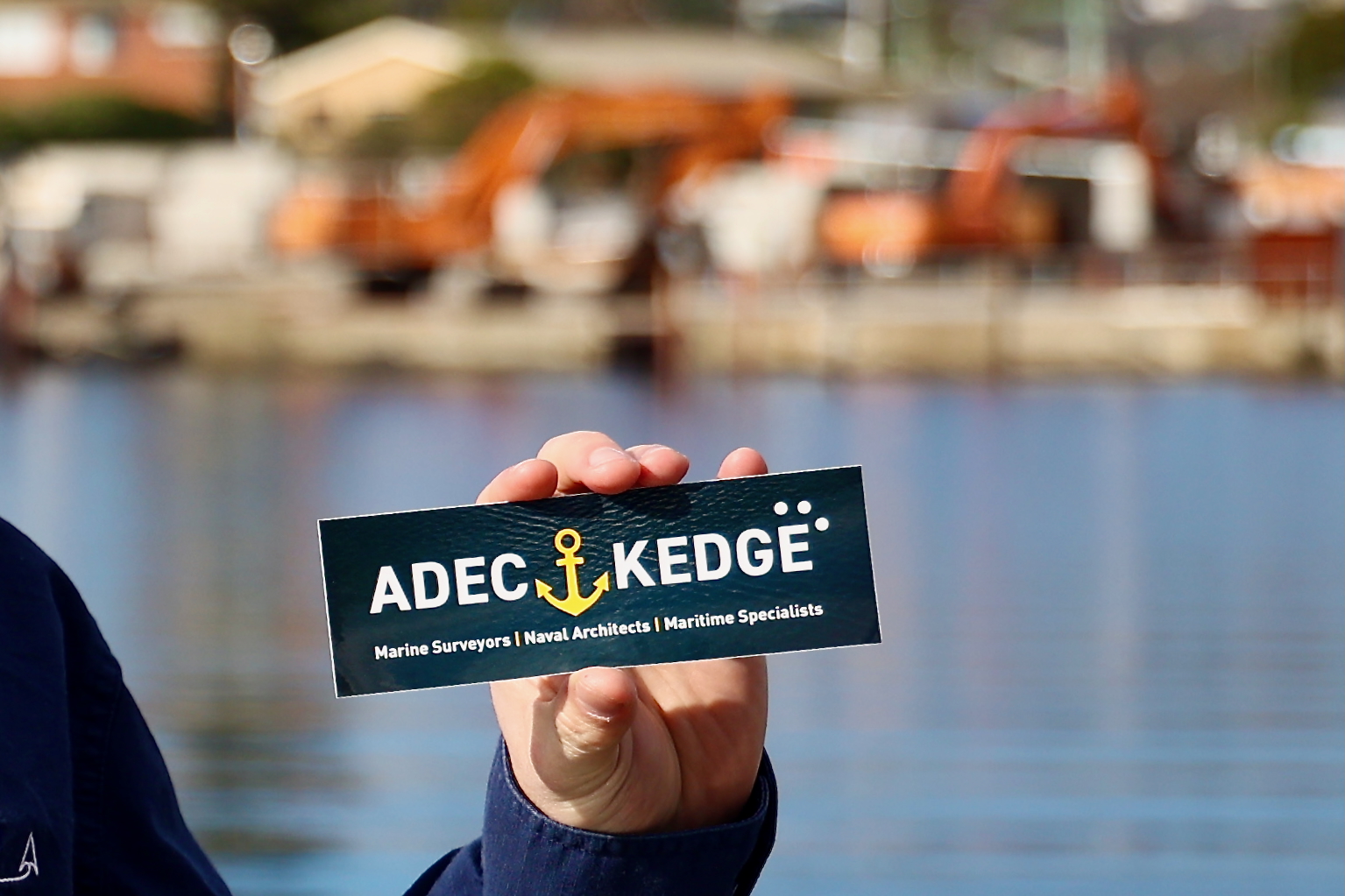 A Hand holding a sticker with the new ADEC Kedge logo featuring the golden anchor.